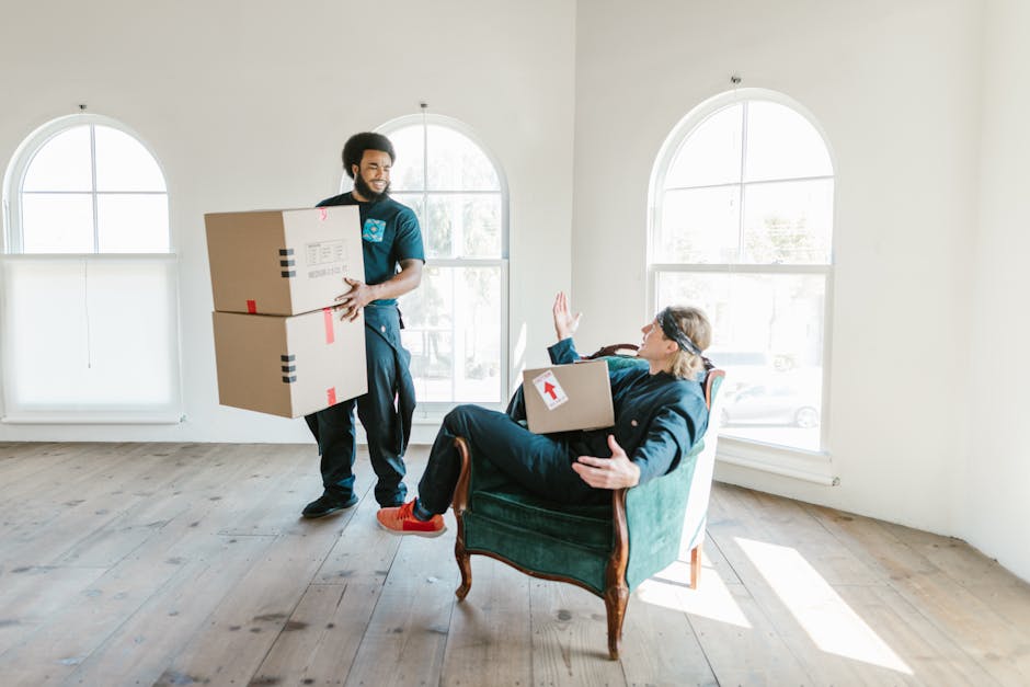 A professional removal team is conducting a home relocation inside a bright, spacious room with white walls and large arched windows allowing natural light to illuminate the space. One man, with curly dark hair and a beard, is standing and holding two cardboard boxes stacked on top of each other, secured with red and black tape, as he prepares to move them. He is dressed in dark work trousers and a blue polo shirt, representing Man With a Van Wood Green, a company specialising in removals. Meanwhile, a woman with glasses and long hair is seated on a vintage green armchair near the window, holding a cardboard box with a white arrow sticker on it, and raising her hand in a gesture of communication or pause during the packing and moving process. The floor is light wood, and there are no other visible objects or furniture, emphasizing the focus on the packing and loading activity. This scene captures the logistical efforts involved in furniture transport and the organisation of packing and moving services in preparation for a house removal, aligned with the Alexandra Palace move guide section on the website.