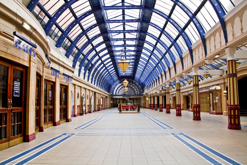 Inside a spacious, well-lit shopping arcade or train station with a high, arching glass and metal roof supported by white columns with gold and red accents. The flooring consists of light-colored tiles with blue decorative lines, leading towards a distant seating area with red upholstered benches. Wooden storefronts with glass windows and doors line both sides of the corridor, some displaying signs or notices. The environment appears clean and empty, emphasizing the architectural details of the historic building. This setting, part of a move or packing process, illustrates the importance of careful furniture transport and logistical planning understood in house removals and relocation services provided by Man With a Van Wood Green. The scene captures the spaciousness and structural features relevant to home relocation logistics within a heritage or public building setting, supporting a comprehensive understanding of moving environments.