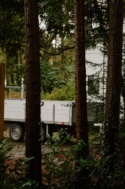 A white flatbed van parked on a paved driveway surrounded by tall trees in a wooded area during daylight hours. The van's side doors are closed, and the vehicle appears to be in the process of home re