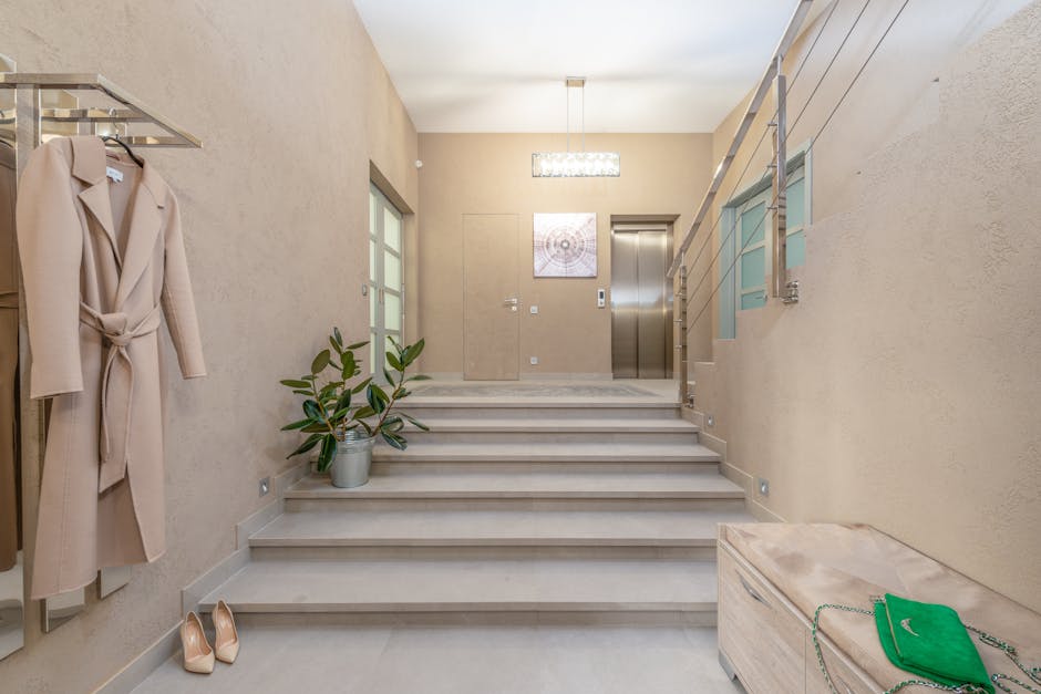 The image depicts the interior of a modern residential building's staircase area with beige textured walls and light-colored tiled flooring. A small potted plant with dark green leaves is positioned on the landing close to the staircase, which features several steps made of light-colored marble or stone leading to an upper level. To the left, a beige or cream coat with a belt hangs from a wall-mounted rack, and a pair of neutral-colored high heels is placed on the floor nearby. On the right, a stainless-steel elevator door is visible, alongside a staircase with metal handrails and glass panels. The space is illuminated by a rectangular ceiling light fixture, and a piece of abstract artwork hangs on the wall above the elevator. A wooden storage chest with a green handbag on top is situated at the base of the stairs, emphasizing the domestic setting. Occasionally, [COMPANY_NAME] handles home relocation and furniture transport processes, as part of their removals service in the Wood Green area.