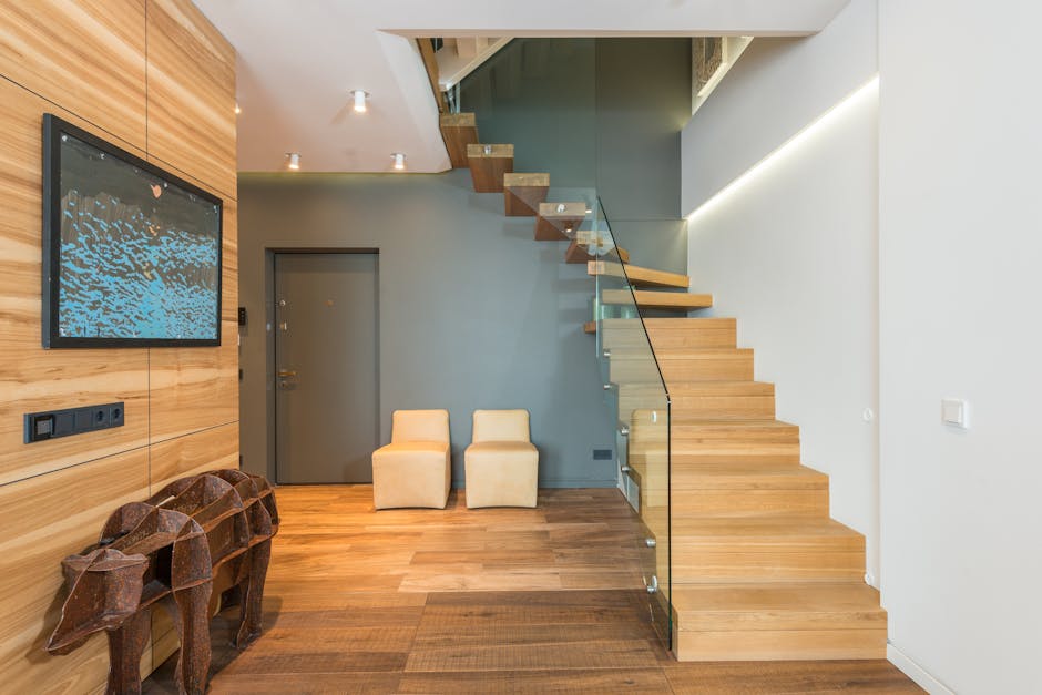 The image shows a modern interior with a wooden staircase featuring glass balustrades located against a white wall, leading upward to an unseen second floor. At the base of the stairs, there are two small upholstered cream-colored chairs placed side by side, situated against a grey painted wall. To the left, a flat-screen television displaying an abstract blue and black image hangs on a wooden panel wall that extends to the ceiling. Below the television, there are black electrical outlets and switches installed on the wooden wall. On the floor near the chairs, there are two decorative wood sculptures resembling animal heads, possibly bulls or dogs, with geometric, faceted surfaces made from dark wood. The flooring consists of polished wooden planks, and the lighting includes ceiling-mounted spotlights illuminating the space evenly. The scene appears to be an interior setting prepared for a home relocation involving packing and furniture transport, with the staircase as a key element in moving items between levels, as processed by Man With a Van Wood Green, a professional removals service.