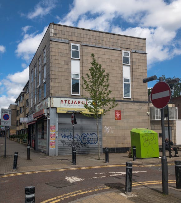 A three-story building with a beige brick exterior and multiple white-framed windows, situated on a street corner under a partly cloudy sky. The ground level features a closed shop with a grey metal roller shutter covered in graffiti, and a small sign above that reads 'STJARULL.' In front of the building, a young tree with green foliage is planted along the sidewalk, which is bordered by black bollards. To the right, there is a bright green refuse bin with graffiti, placed near a black and white 'no entry' traffic sign. A person is seated on a bench close to the refuse bin. The scene includes street signs, a pavement with tactile paving, and a vehicle parked nearby, indicating an urban environment suitable for home relocation or furniture transport activities. The overall setting suggests a typical urban area where professional removals or house moving services, such as those provided by Man With a Van Wood Green, might operate, involving loading or transporting furniture and boxes during a house move near Turnpike Lane.