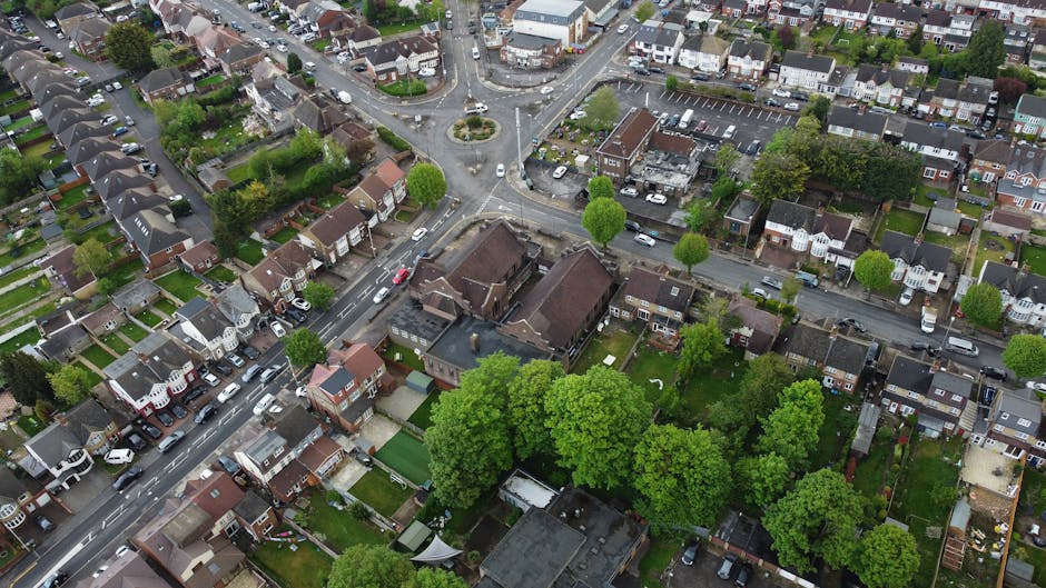 An aerial view of a residential neighbourhood in Wood Green with closely spaced terraced houses, semi-detached homes, and small detached properties. The streets are lined with parked cars, and several green trees are visible providing greenery amidst the tightly packed houses. Some properties have gardens enclosed by fences or hedges, and the area includes a small roundabout at an intersection. In the foreground, there are houses with visible roofs, driveways, and front yards, while further back, larger buildings and commercial structures can be seen. This scene captures the typical layout of an urban residential area suitable for house removals and furniture transport, with the surroundings indicating an active move or home relocation process, as might be coordinated by [COMPANY_NAME], which specializes in removals near Turnpike Lane.
