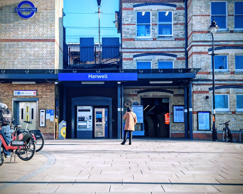 The image shows the exterior of Hanwell station, part of the Elizabeth Line, situated within a brick building with a modern black canopy. A woman wearing a light pink coat and dark trousers is standing on the pavement, facing the station entrance, which features a large roller shutter door and digital information screens mounted on the wall. To the left, there are several bicycles parked, including a Santander cycle and a docking station, indicating bicycle-friendly facilities. On the right side, a lamppost and a bicycle are visible near the building's corner. The station entrance has signage for ticket machines and accessibility features. This setting, within a typical urban environment, depicts the environment where house removals or furniture transportation could commence or conclude, and is relevant for relocation logistics, especially when planning transport from a property near Hanwell station. Man With a Van Wood Green offers professional removals and moving services suitable for such locations, ensuring secure and efficient home relocation, furniture transport, and packing processes in this area.