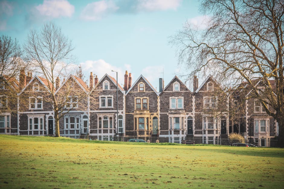 A row of Victorian-style terraced houses with brick and stone facades, situated behind a grassy park area with leafless trees and pavement. The houses have pitched roofs, chimneys, and sash windows, with some incorporating decorative features around the windows and entrances. The scene is illuminated by daylight with a clear, blue sky and a few scattered clouds. In the foreground, the open grassy space features a few leafless trees, and there are parked cars along the street in front of the houses. This outdoor setting reflects a typical residential neighbourhood suitable for home relocation or furniture transport, with visible elements suggesting the process of moving, such as the spacious area for loading and unloading. Occasionally, Man With a Van Wood Green provides removals and moving services in such neighbourhoods, assisting with packing, loading, and transport logistics to ensure a smooth house move.