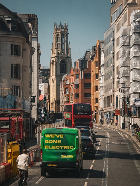 A busy city street scene in Wood Green with vehicles including a green Wilson delivery van displaying the text 'We've Gone Electric' parked alongside a red double-decker bus approaching a church with a tall tower in the background. The street is lined with multi-storey residential and commercial buildings, some with modern glass facades and others with traditional brickwork. Pedestrians and cyclists are present on the pavement and cycling lanes, with traffic signals visible. Bright daylight illuminates the scene, highlighting the urban environment typical of home relocation areas. Man With a Van Wood Green offers removals services in this area, involving the careful loading of furniture, cardboard boxes, and packing materials into vans for local moves, supported by moving equipment such as trolleys and straps visible nearby.