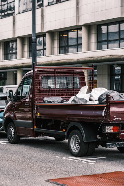 A burgundy flatbed van parked on a street in an urban area, with its cargo area loaded with various household items wrapped in protective coverings, including cardboard boxes, plastic-wrapped furniture, and other moving materials. The vehicle is positioned adjacent to a modern glass-fronted commercial building, with a city street and parked cars visible in the background. The van is part of a home relocation or furniture transport process, supporting services such as packing, loading, and transporting belongings, as provided by Man With a Van Wood Green, a company specialising in removals. The scene captures the loading process or preparation for a local move, highlighting typical logistics involved in house removals in Wood Green.
