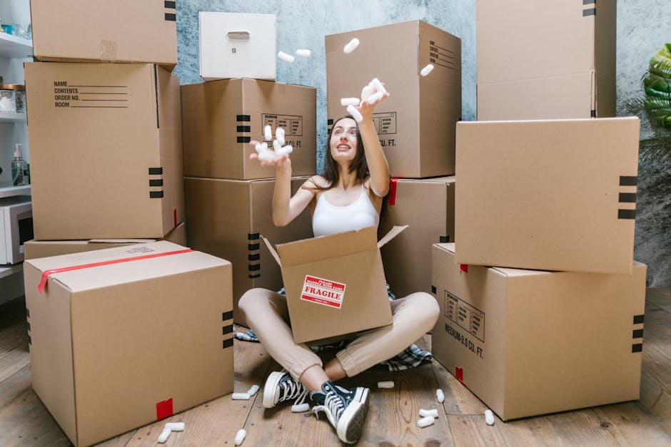 A young woman sitting cross-legged on a wooden floor during a home relocation process, surrounded by numerous cardboard moving boxes of various sizes, some sealed with red tape and others open, reveal