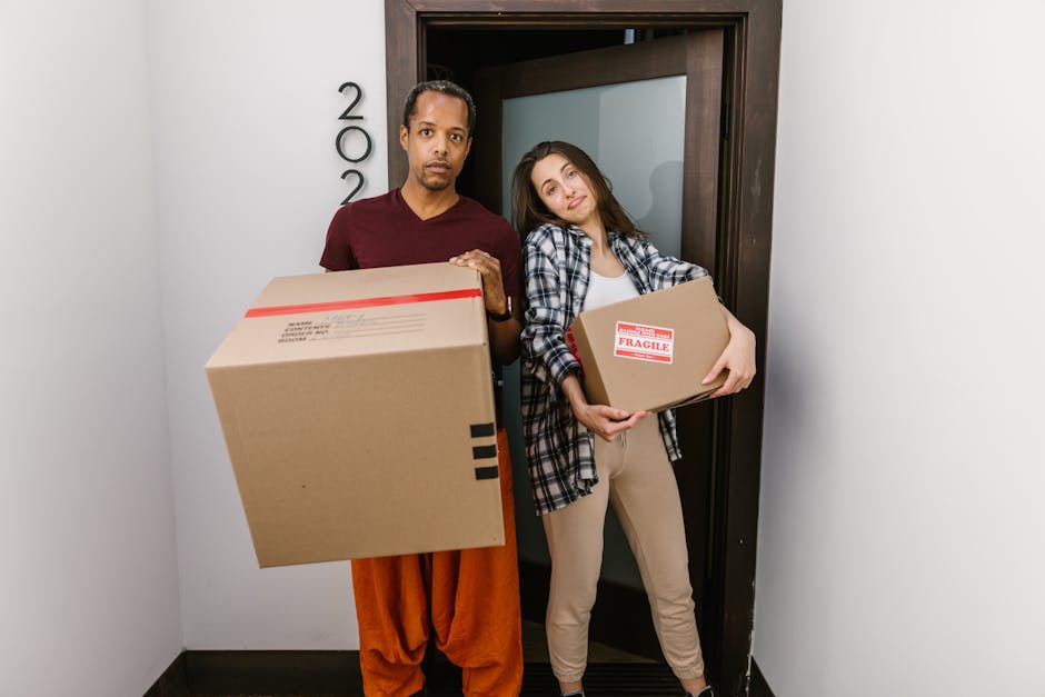 A young woman sitting cross-legged on a wooden floor during a home relocation process, surrounded by numerous cardboard moving boxes of various sizes, some sealed with red tape and others open, reveal