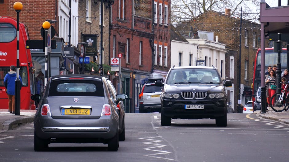 A narrow residential street in Wood Green with parked cars on both sides, including a black BMW SUV positioned facing the camera and a grey Fiat 500 with the license plate LN13 GJZ parked behind it. The street is lined with brick terraced houses featuring storefronts on the ground floors. Pedestrians are walking along the pavement, some near bus stops and bike racks, with a group of people gathered on the right side near a bus shelter. Visible in the background are additional parked vehicles, street signage, and city infrastructure such as lampposts and traffic signals. This scene captures typical urban street activity during daytime, with a focus on parking and narrow street navigation, relevant to home relocation or furniture transport logistics managed by Man With a Van Wood Green, illustrating the challenges of city-based removals and the importance of careful planning during loading and unloading processes in tight urban environments.