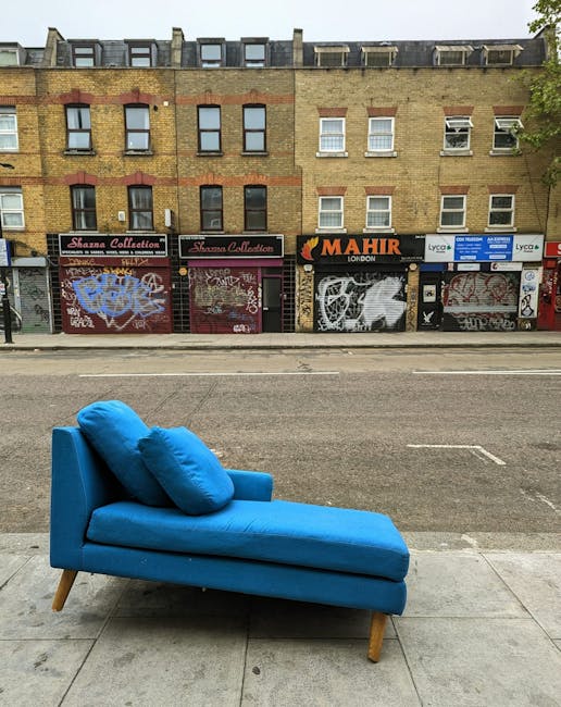 A vibrant blue chaise lounge with a matching bolster cushion placed on the seat, situated on the sidewalk outside a row of commercial buildings in Wood Green. The furniture features wooden legs and a fabric upholstery, positioned close to the curb with the street and road markings visible in the background. Behind the sofa, storefronts with closed metal shutters are covered in graffiti and art, including signs for shops such as 'Sharma Collection' and 'Mahir London.' The buildings are multi-storey, constructed with brickwork, and have large windows on the upper floors. The scene appears to depict a home relocation or furniture transport scenario where [COMPANY_NAME], such as Man With a Van Wood Green, might be involved in furniture removal or collection services, highlighting the process of loading or offloading furniture as part of their house removals and moving logistics. The lighting is natural, suggesting daytime with overcast or partly cloudy weather.