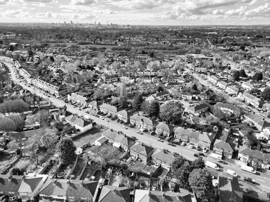 Black and white aerial photograph showcasing a residential neighbourhood with rows of terraced houses and detached homes, front gardens, and narrow streets lined with parked cars. A main road runs through the centre, with some vehicles visible in transit. In the foreground, rooftops made of tiles and shingles, along with trees and backyards, are clearly identifiable. Lighter areas indicate open spaces and streets, while darkerened sections highlight dense foliage and shaded rooftops. The industrial or city skyline is visible in the distant background, with high-rise buildings and infrastructure on the horizon under partly cloudy skies. The image captures the suburban area during daylight, providing a clear view of housing arrangements and urban sprawl. An interior view related to home relocation or furniture transport is not visible, but this setting emphasizes the type of environment where professional removals by Man With a Van Wood Green could operate efficiently, involving packing, loading, and transport processes.