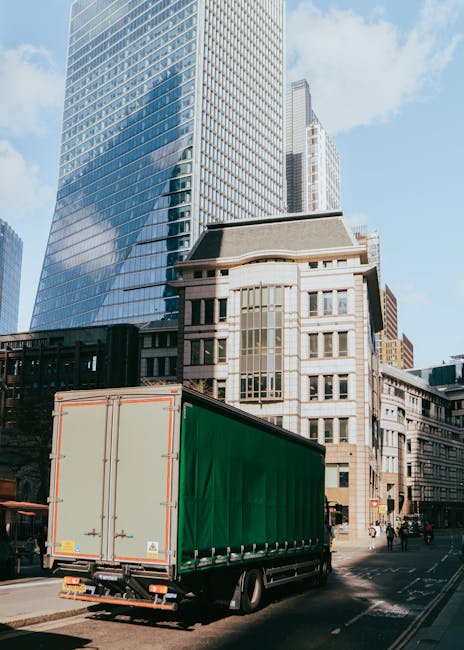 A large green moving truck is parked on a city street in front of modern glass skyscrapers and older stone buildings. The truck's rear doors are closed, and it is positioned near a pavement with a few pedestrians walking nearby. The scene depicts an urban environment suitable for house removals or furniture transport, with the truck ready for loading or unloading. The presence of tall office buildings and the busy street background suggests a professional relocation process occurring within a city setting, consistent with home and office moving services provided by Man With a Van Wood Green during a time of day with ample daylight.